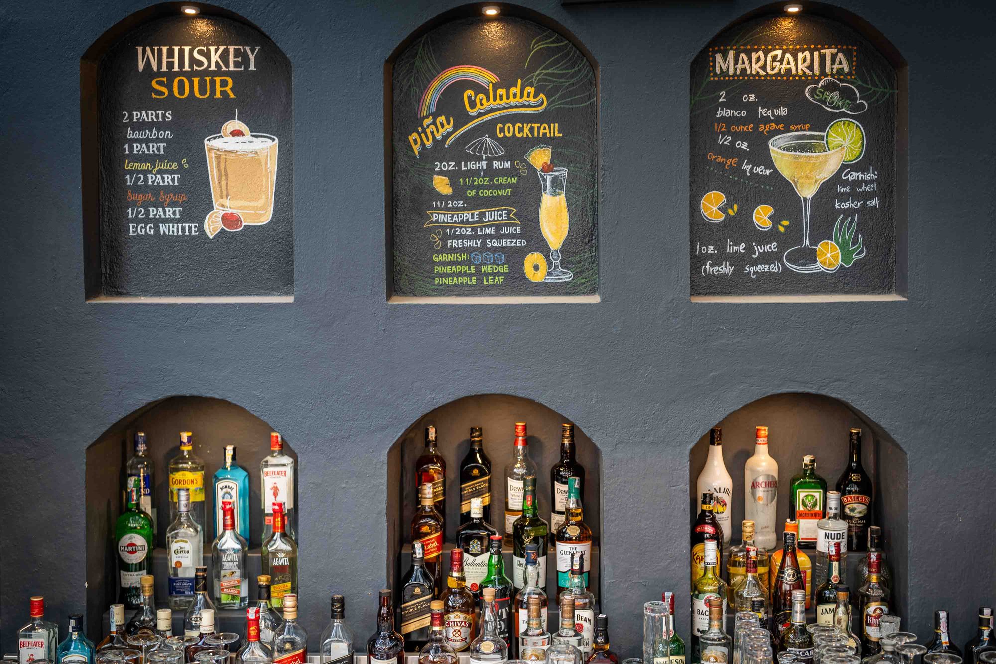 Hand-chalked whiskey sour, piña colada, and margarita recipes above the lit spirits niches at Jharana Bar